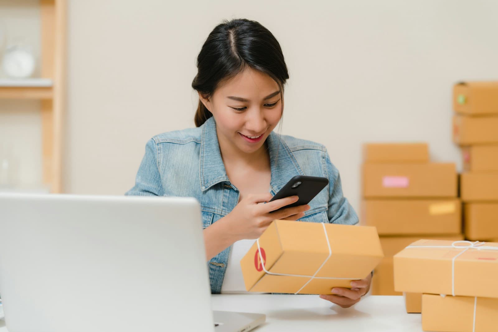 A person preparing packages while checking a phone beside a laptop.